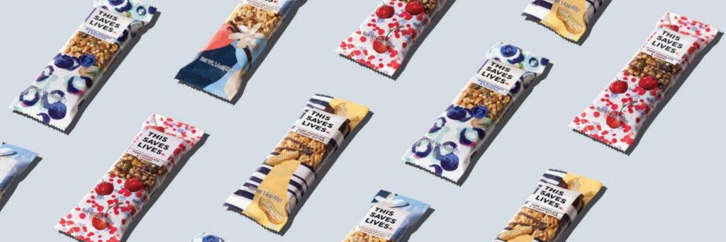An image of a variety of snack bars neatly arranged in rows on a white surface. The snack bars are individually wrapped and come in different flavors. The packaging features vibrant colors and the logo of 'This Saves Lives' prominently displayed on each wrapper.