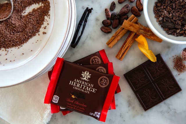 A square of dark chocolate surrounded by a white plate dusted with cocoa powder and garnished with delicate strips of orange peel and cinnamon sticks, arranged on a marble table