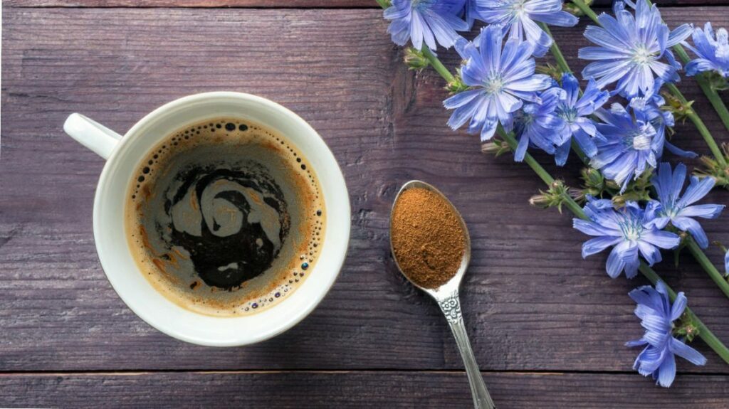 A white mug filled with chicory root coffee next to a silver spoon with chicory root coffee grounds, adorned with blue flowers, on a wooden table.