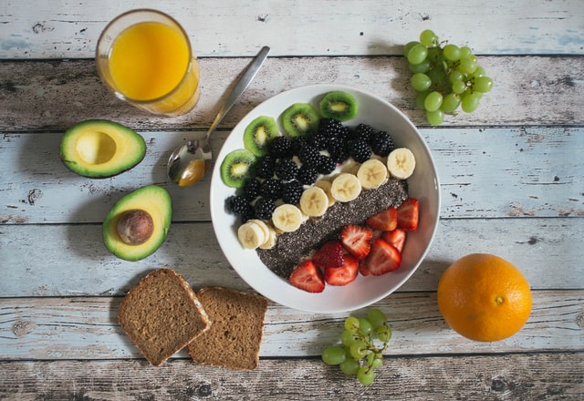 An acai bowl filled with vibrant purple acai puree topped with sliced strawberries, bananas, granola, chia seeds, blackberries, and kiwi, served in a white bowl on a wooden surface surrounded by an orange, avocado, grapes, and a glass of orange juice.