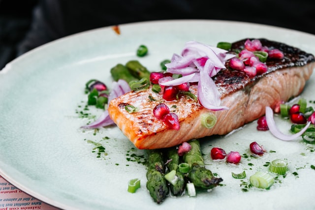 A grilled salmon fillet garnished with thinly sliced red onions and vibrant pomegranate seeds, accompanied by roasted asparagus spears on a white plate