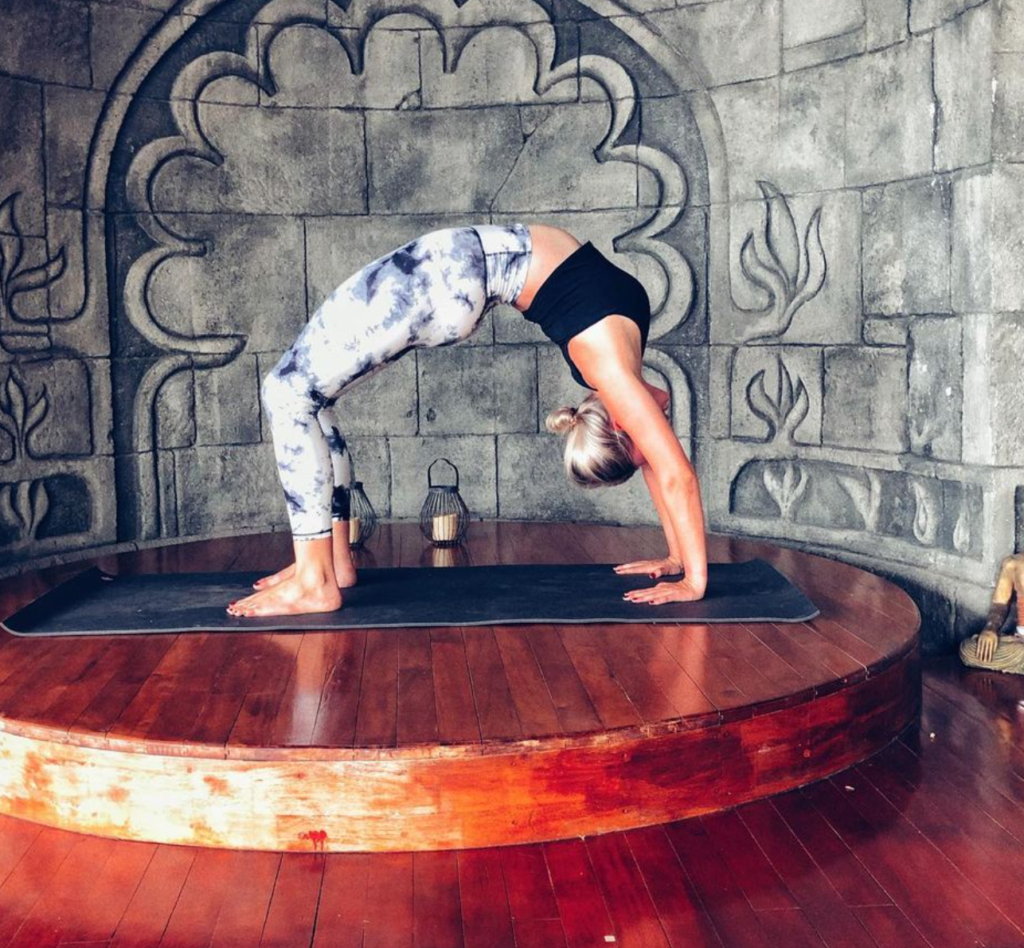 A woman wearing tie-dye leggings and a black top performing a backbend on a grey yoga mat placed on a wooden stage.