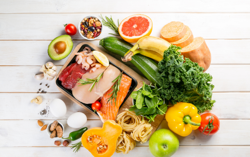 Assorted foods arranged in a circle on a white shiplap background. Includes salmon, avocado, pasta, bananas, nuts, grapefruit, tomatoes, garlic, zucchini, sweet potatoes, yellow bell peppers, kale, squash, jalapenos, green apple, spinach, eggs, chicken breast, beef, shrimp, lemon, and herbs.