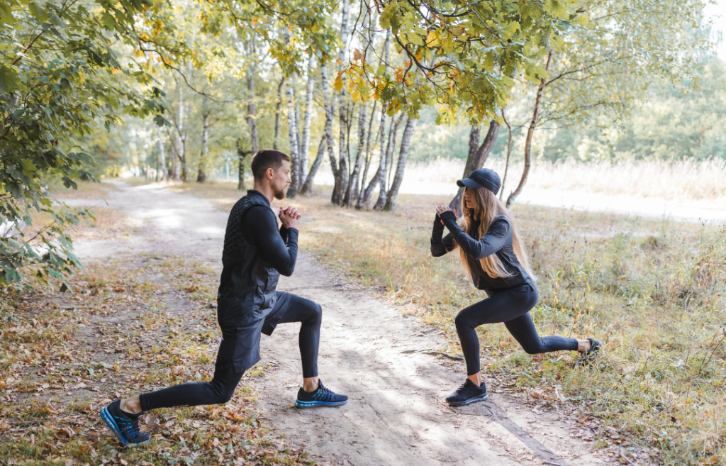A man and a woman wearing all-black fitness clothing performing lunges on a nature trail.