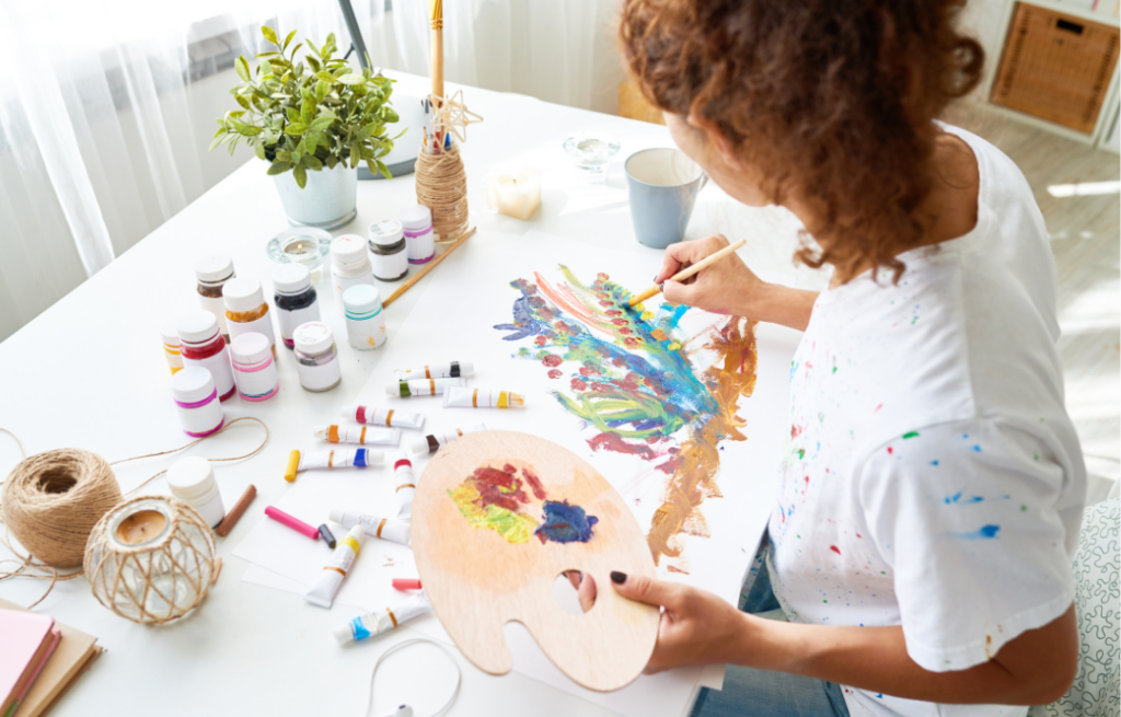 Focused woman painting on a white table, surrounded by an array of colorful paint tubes and a wooden palette with mixed paints