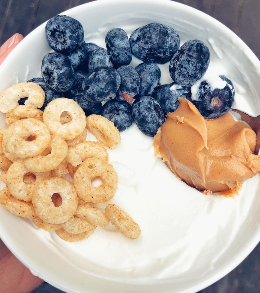 A white bowl filled with yogurt topped with fresh blueberries, Cheerios cereal, and a drizzle of creamy peanut butter.