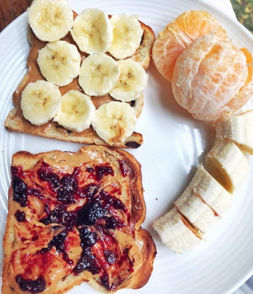 Peanut butter banana toast on whole wheat bread beside another slice topped with peanut butter and jelly, accompanied by a peeled clementine and sliced banana.