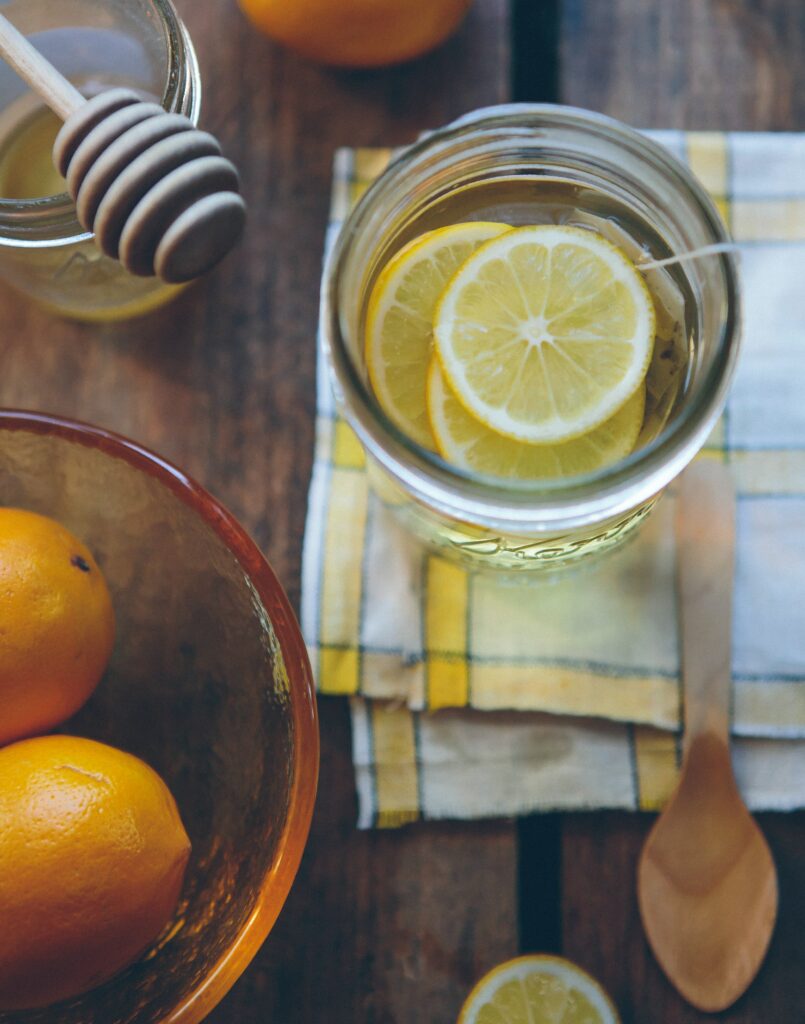 Mason jar filled with hot water and lemon slices steeping tea on top of a yellow plaid kitchen cloth, alongside a honey mason jar with a dipper and a glass bowl containing fresh lemons. 