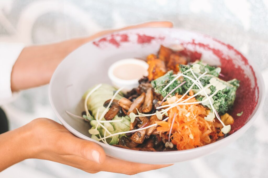 Woman holding a bowl filled with greens, roasted sweet potatoes, grilled chicken, and tangy kimchi. 