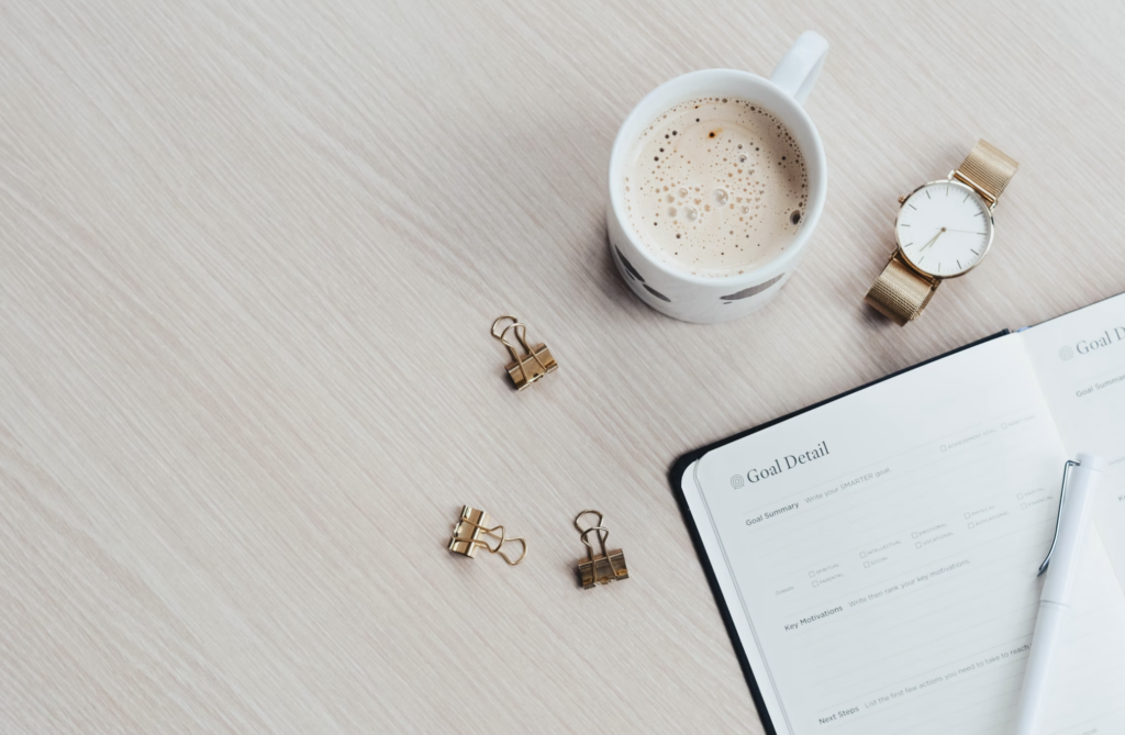 Cup of coffee and goals journal on a white table next to gold paper clips and a watch